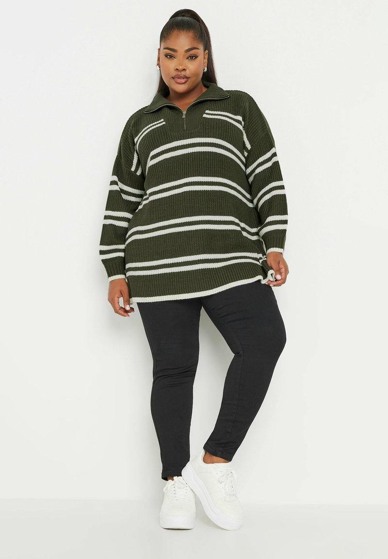 Stickad jumper i mörkgrön med vita ränder, med hög krage och dragkedja. Kombinerad med svarta byxor och vita sneakers.