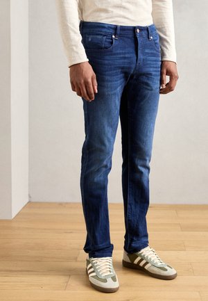 Mężczyzna ubrany w dopasowane niebieskie jeansy, beżową koszulę z długim rękawem oraz oliwkowo-białe trampki stojący na drewnianej podłodze.