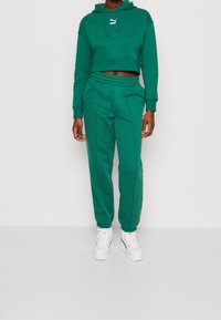 Sweat à capuche vert crop avec cordons et logo, associé à un pantalon de jogging assorti. Les deux sont fabriqués dans un tissu doux avec des poignets et une taille élastiques.