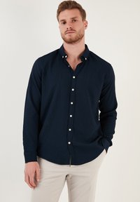 Camisa de botones azul marino de manga larga con tejido texturizado, pequeño cuello y botones blancos, combinada con pantalones de color claro.