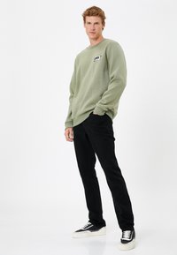 Sudadera verde oliva con cuello redondo y un pequeño gráfico. Combinada con pantalones negros ajustados y zapatillas en blanco y negro.