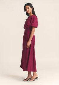 Mauve keskikokoinen mekko, jossa on puffipäät lyhyet hihat, tiukka yläosa ja haihtuva hame, yhdistettynä mustiin flip-flop -sandaaleihin. Tasainen kankaan rakenne.