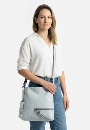 Frau mit schulterlangem blondem Haar, die ein weißes Hemd und blaue Jeans trägt und eine hellgraue Umhängetasche mit Quasten-Detail hält.