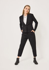 Completo nero su misura con giacca aderente a un bottone, abbinato a pantaloni slim-fit e stivali alla caviglia. Tessuto morbido, design minimale.