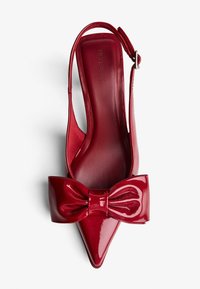 Rdeče lakaste slingback čeveljčke s koničastim nosom in velikim vozelom. Gladka tekstura z bleščečim videzom. Znamka vtisnjena znotraj.