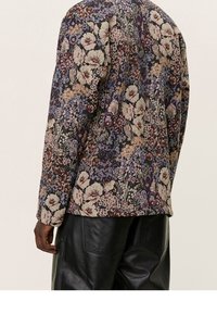 Blazer con estampado floral en tonos oscuros, tejido texturizado y corte estructurado, combinado con pantalones sueltos de cuero negro.