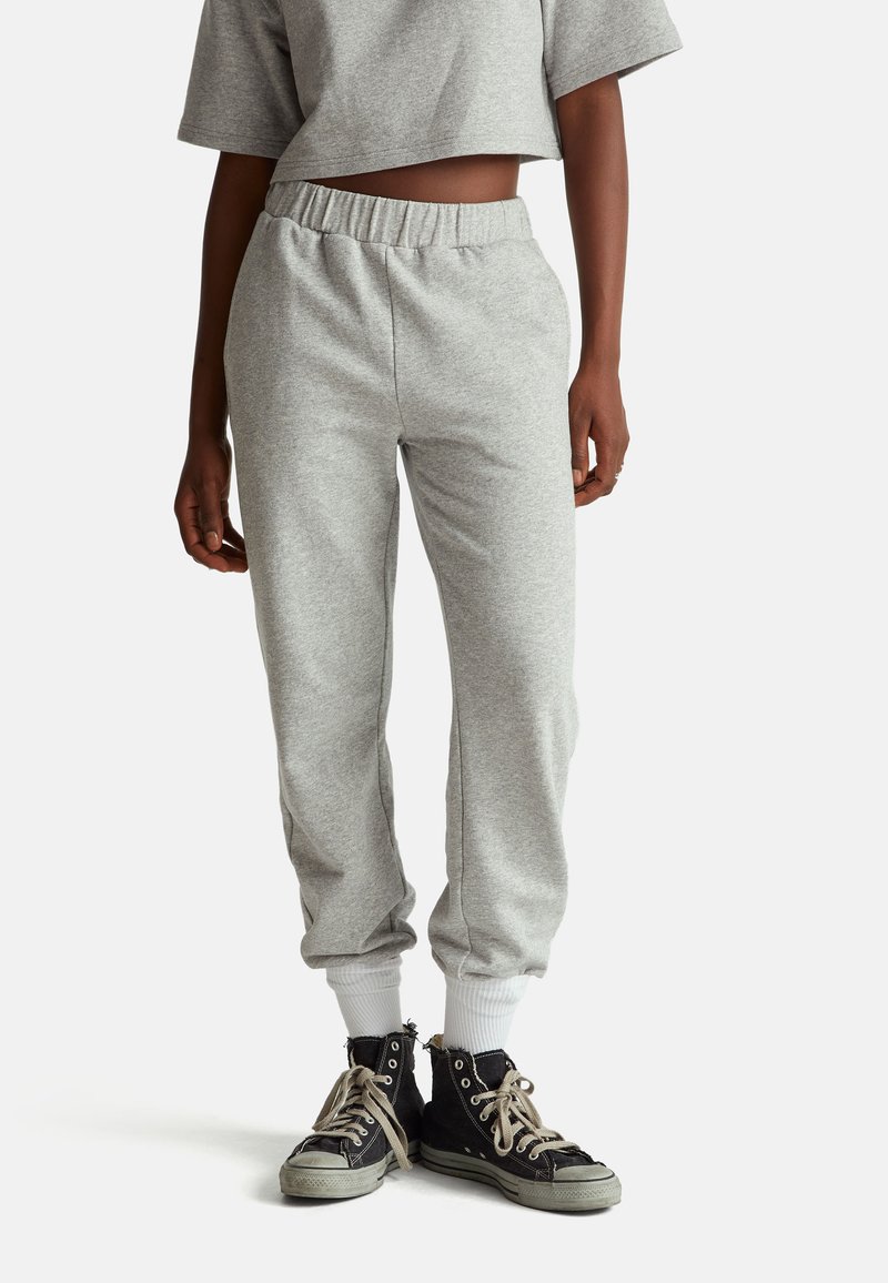 Šedé tepláky s elastickým pasem, žebrovanými manžetami a měkkou texturou, v kombinaci s černými kotníkovými teniskami s béžovými tkaničkami.