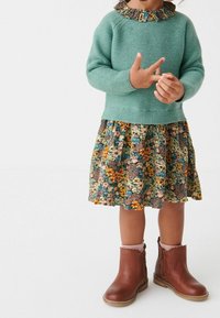 Pletený zelený svetr s volánkovým květinovým límcem, kombinovaný s šaty s květinovým potiskem a sklady v odstínech oranžové a modré, hnědé kotníkové boty.