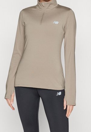 Kobieta ubrana w beżową bluzę z długim rękawem i półzamkiem oraz czarne legginsy, oba z białymi logo New Balance, stojąca na jednolitym tle.