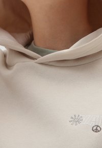 Béžová mikina s kapucí, která vyniká měkkou texturou, vyšívanými detaily slunce a symbolu míru, a vrstveným zeleným límcem.