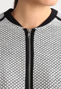 Vit och svart zip-up jacka med ett texturerat mönster. Har en rund halsringning och svarta detaljer längs med dragkedjan och ärmhålen.