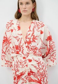 Witte jurk met rood bloemenpatroon, diepe V-hals en gathered taille. Heeft brede kimono-mouwen en decoratieve rode oorbellen.