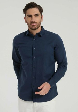 Homme portant une chemise à boutons bleu foncé et un pantalon blanc, debout devant un fond clair uni, ajustant sa chemise d'une main.