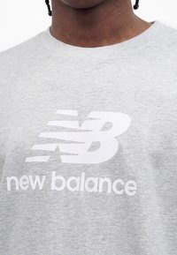 Close-up van een grijs shirt met ronde hals en een wit New Balance-logo en tekst, gecentreerd op de borst.