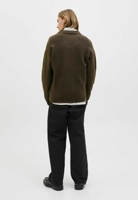 Person with shoulder-length blond hair wears brown sweater, white shirt, black pants, and black shoes, standing facing away against white background.