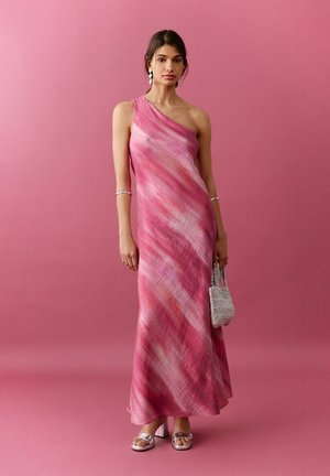 Donna che indossa un abito lungo monospalla rosa e rosso, sandali argento, con in mano una piccola borsa grigia intrecciata, su uno sfondo rosa uniforme.