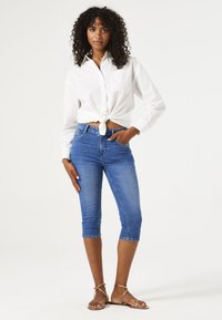 Witte blouse met knopen, geknoopt in de taille, gecombineerd met knielange blauwe spijkerbroek. Gouden sandaaltjes met bandjes maken de look compleet.