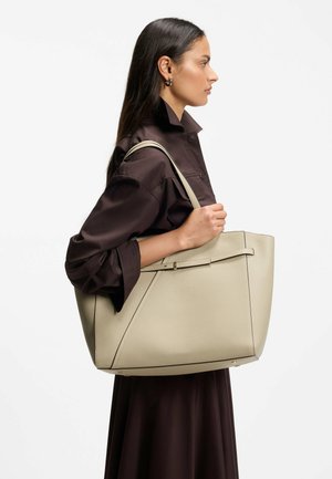 Woman in a dark brown coat holding a large beige leather tote bag over her shoulder against a plain white background.