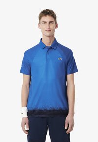 Blå pikétröja med en gradientdesign som går från klarblå till mörkare blå vid fållen, korta ärmar och en Lacoste-logotyp på bröstet.