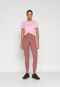 Ροζ κοντομάνικη μπλούζα με σχεδιασμό εμπρός με στρίψιμο, συνδυασμένη με ψηλόμεσα κολάν σε σκουρόχρωμο κεραμιδί. Το μοντέλο φοράει μαύρα αθλητικά παπούτσια.