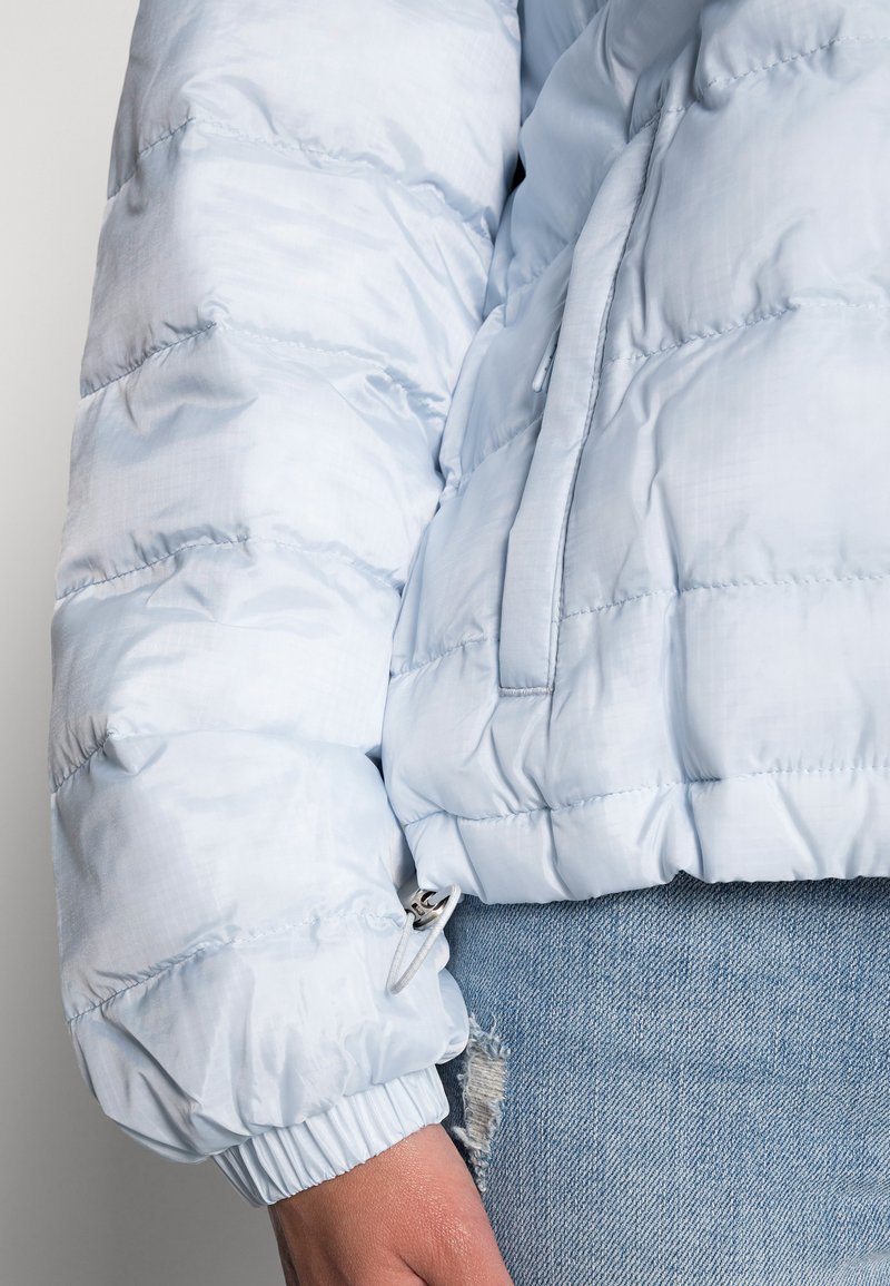 Giacca puffer azzurro chiaro con texture trapuntata, polsini elastici e tasca laterale. Presenta una finitura liscia e lucida e un materiale morbido.