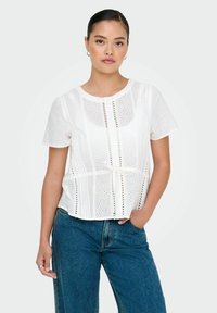 Weiße Bluse mit kurzen Ärmeln, die dekorative Lochdetails und einen Knoten in der Taille aufweist, kombiniert mit blauen Jeans aus Denim. Weiche Textur.