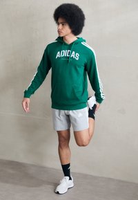 Grön hooded tröja med vit logotyp och ränder, grå shorts, svarta strumpor och vita sportskor. Modell som sträcker ut sig mot en enkel bakgrund.