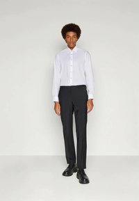 Weißes Hemd mit langen Ärmeln, kombiniert mit schwarzer Anzughose. Das Model trägt schwarze Schuhe und steht vor einem neutralen Hintergrund.