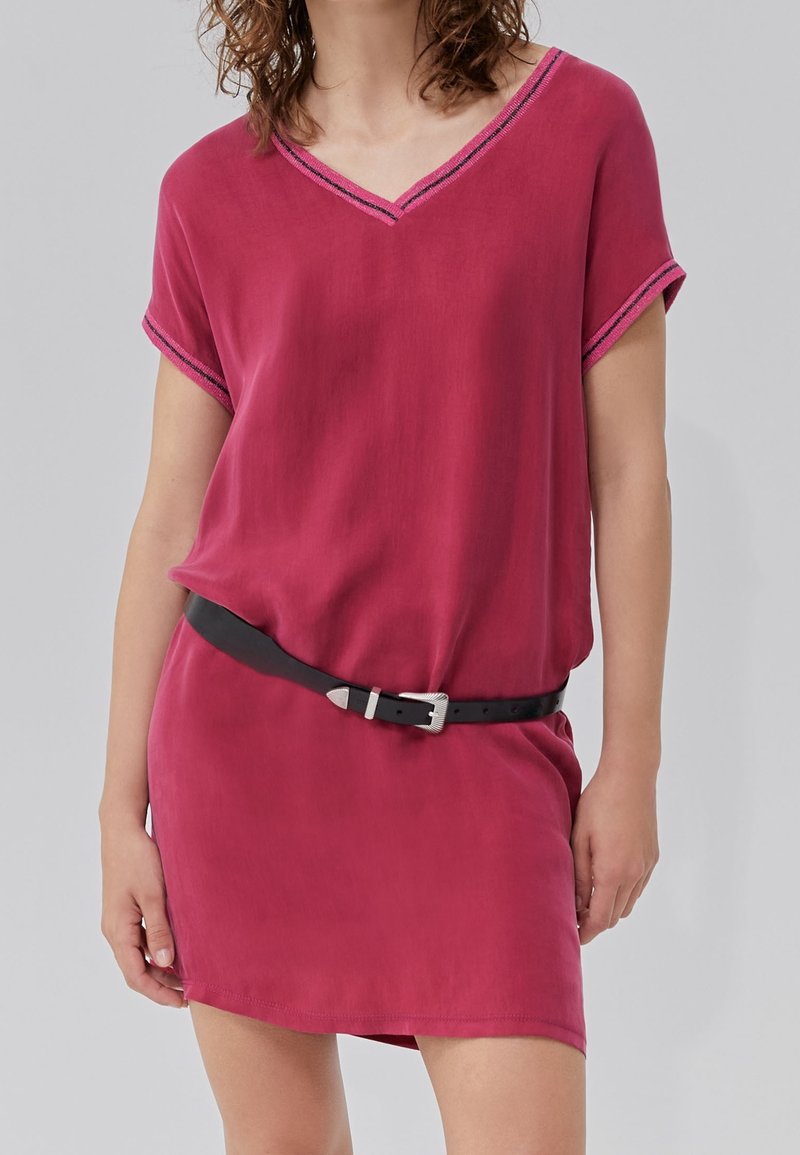 Femme portant une robe rose foncé à manches courtes avec un bord noir aux manches et à l'encolure, stylisée avec une ceinture en cuir noire à la taille.