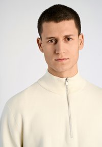 Krämfärgad zip-up tröja med hög krage, tillverkad av mjukt tyg. Har en silverdragkedja framtill. Slät textur, minimalistisk design.