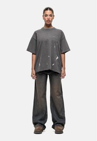 Szara, oversize'owa koszulka z logo na przodzie, z detalami w postaci plam farby. Połączona z luźnymi, ciemnoniebieskimi dżinsami i brązowym obuwiem.