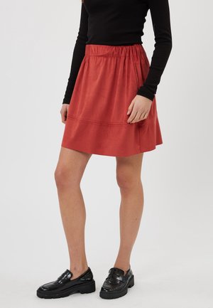 Frau trägt ein schwarzes Langarmshirt, einen roten Rock mit elastischem Bund und schwarze klobige Loafer, steht vor einem schlichten Hintergrund.