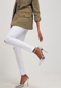 Chaqueta de utilitario verde oliva, jeans ajustados blancos con detalles rasgados y zapatos de tacón alto plateados brillantes con correas en los tobillos.