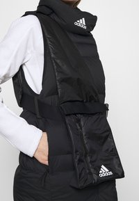 Sort polstret vest med høj krave og justerbare remme. Har en lynlåslomme og Adidas-logoet på forsiden. Blød tekstur.