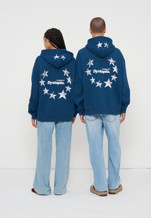Deux personnes portent des sweat-shirts à capuche bleu marine avec des étoiles blanches usées formant un cercle et le texte "unspoken Dystopia" dans le dos, associés à un jean bleu clair.