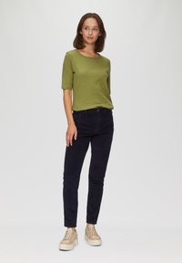 Oliwkowa, prążkowana bluzka z krótkim rękawem w połączeniu z ciemnoniebieskimi spodniami z korduroy i beżowymi trampkami o fakturowanej podeszwie. Model stoi na jasnym tle.