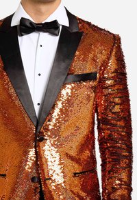 Chaqueta de esmoquin de lentejuelas con lentejuelas naranjas y doradas. Presenta solapas de satén negro y ribete en los bolsillos, y una camisa blanca con corbata de moño negra.