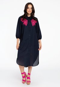 Navyblauwe shirtjurk met geborduurde rozenkwartsaccenten, knoopsluiting en driekwart pofmouwen. Gedragen met roze wedge-sandalen.