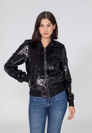 Vrouw met donker haar draagt een zwarte pailletten jas met rits en blauwe jeans, staande tegen een effen lichtgrijze achtergrond.