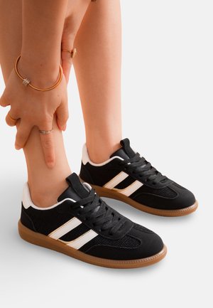 Personne portant des baskets noires à rayures blanches et semelles en gomme, tenant une cheville avec les deux mains ornées de bagues et de bracelets.