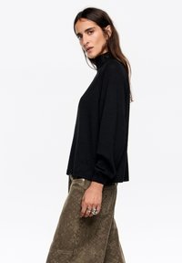 Czarny dzianinowy sweter z wysokim kołnierzem, luźnym krojem i długimi rękawami. Noszony z oliwkowymi spodniami o fakturze i srebrnymi pierścionkami na palcach.