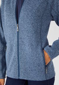 Chaqueta de forro polar azul con textura jaspeada, cremallera de longitud completa y bolsillos laterales. Presenta tejido en contraste alrededor de la cremallera y los puños.