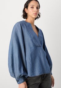 Denimbluse i middels blå med dyp V-hals, lange, vide ermer og knappe mansjetter. Har en avslappet passform og subtil tekstur.