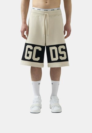 Mężczyzna w beżowych krótkich spodenkach do kolan z czarnymi panelami i dużym napisem "GCDS", białe skarpetki i białe trampki na rzepy.