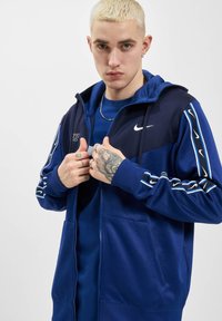 Jūras spēku krāsas puloveris ar rāvējslēdzēju un baltu Nike logo, sānu svītrām un kabatām kā kangars. Iekļauj ribotas piedurknes un kapuci ar auklu.