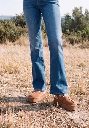 Une personne portant un jean bleu et des pantoufles marron en daim à plateforme se tient sur un sol sec dans un champ herbeux.