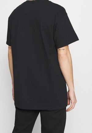 Person wearing a plain black short-sleeve t-shirt and black pants, viewed from the back, showing barbed wire tattoos on both arms.