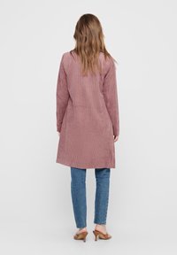 Langer, gerippter, rosafarbener Mantel mit geradem Schnitt und seitlichen Schlitzen, kombiniert mit blauen Jeans und hellbraunen Mules mit Absatz.