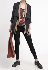 Veste noire drapée sur un cardigan côtelé marron, haut à fleurs, jean skinny noir et chaussures noires à lacets. Sac à main marron avec une finition texturée.