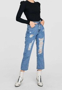 Kvinna iklädd svart långärmad åtsittande topp, hög midja trasiga blå jeans och vita metalliska ankelstövlar, står med händerna i fickorna.